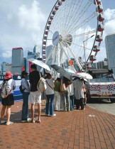 السائحون يستخدمون المظلات لحماية أنفسهم من أشعة الشمس أثناء اصطفافهم لشراء الآيس كريم في ميناء فيكتوريا بهونغ كونغ. (ا ف ب)